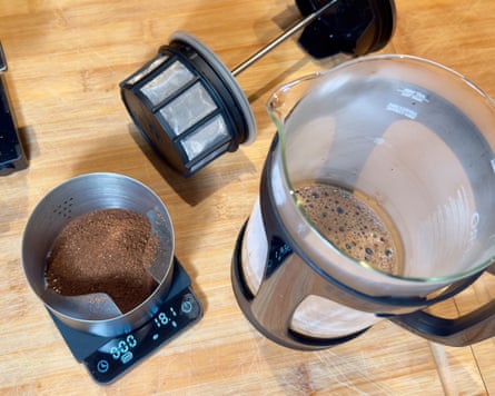 A cafetiere with coffee in it on a wooden worktop surrounded by other coffee paraphernalia