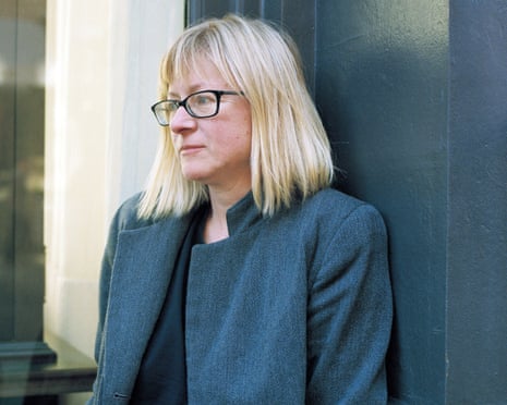 Helen DeWitt, wearing glasses and a jacket, leans against a wall looking away