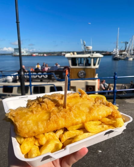 Hake and chips in a polystyrene tray, with boats in a harbour in the background.
