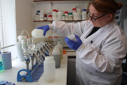 A woman wearing a white laboratory coat and latex gloves pours liquid from a bottle into one of a row of transparent containers