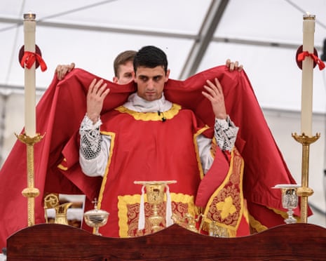 A priest in ornate red vestments is helped to put on ceremonial robes over an altar