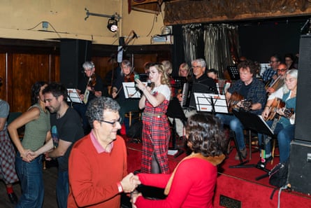 An older man dances with a woman by a younger couple in front of the stage and musicians. A young blond woman in a tartan dress is calling the dance moves.