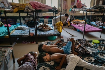 A woman sweeps between metal bunk beds, while women and children sleep on some of the beds, on the floor and on a mattress.