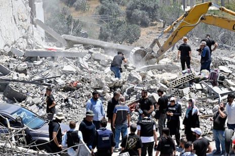 A small crowd of people gather under a bright sun before the white-grey rubble of a destroyed building, the arm of a yellow excavator sending up a cloud of white-grey dust behind them.