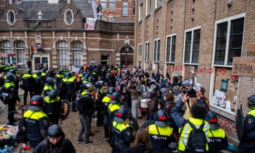 a large group of riot police wearing helmets and high-vis jackets surround a group of protesters in a courtyard at the university, which is a historic brick building. One student is seen on the roof with a banner and some are holding cardboard placards; there is graffiti scrawled in red paint on the walls.