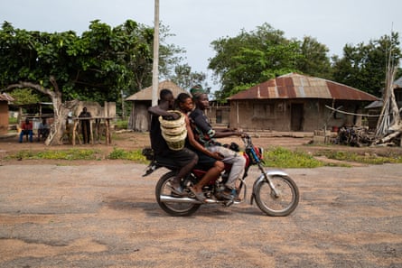 Three men on a motorbike pass mud-brick houses and a group of men sitting in the shade of a tree.