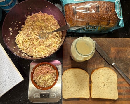 An overhead photo of a chopping board with a loaf of bread, two slices of bread, an open jar of mayonnaise and a bowl of cheese and tuna mix with a smaller bowl of the mix on digital scales.