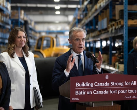 A man at a speaks into a microphone at a lectern that says 'Building Canada Strong for All' in English and French as a smiling woman stands behind him.