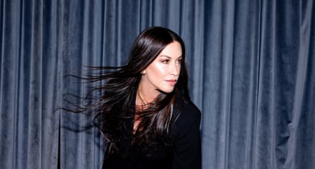 Portrait of Alanis Morissette in a dark jacket against a dark curtain with her shiny dark hair billowing about her