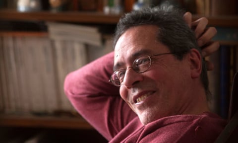 Cesar Aira, smiling at the camera, scratches the back of his head. Behind him are some books on a shelf.