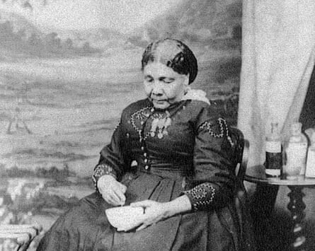 Mary Seacole sits in a chair in a long dark dress, holding a small bowl. There is a table beside her holding bottles of what may be medicines.