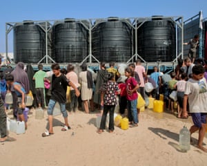 People queue up with jerry cans next to large black plastic water tanks