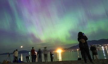People stand on a beach at night looking towards the northern lights in the sky above the water. The lights have coloured the sky purple and green