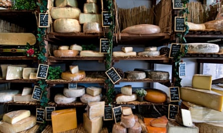 Cheeses of different sizes and colours sit on shelves with name and price labels beside them