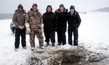 Five people in winter gear stand in a row on a snowy beach with a waterlogged piece of wood in front of them.