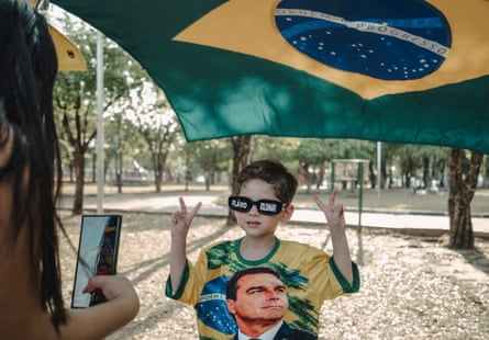 Fábio Heitor poses making peace signs with his hands under a Brazilian flag, wearing a Bolsonaro shirt, as his mother takes a picture with her smartphone