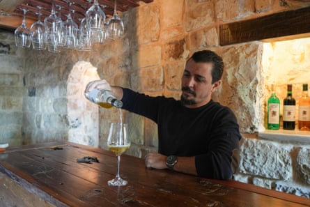 Shadi Jarjour pours a glass of wine on a wooden counter; wine glasses hang from a shelf above him and there are bottles of wine in an illuminated alcove in the stone wall behind him. He has slicked-back black hair and a neat pointed beard with a few grey strands, and wears a black jumper with brown elbow patches and a large wristwatch.