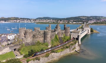 The castle has rounded turrets, and a train is snaking past it, having crossed the bridge over the bay.