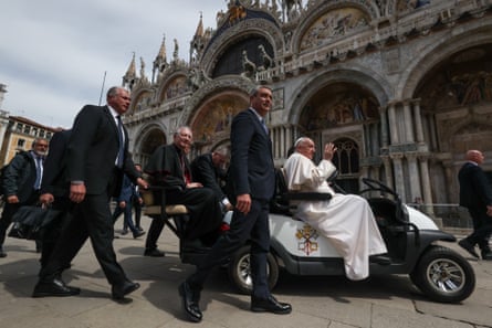 Pope Francis waves as he is driven in a golf cart-like vehicle through a square; men in dark suits walk alongside the cart