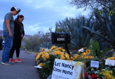 A man and woman look down at a memorial of flowers and a sign that says “Let Nancy come home” next to a driveway mailbox.