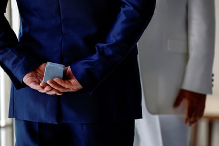 A male couple wearing blue and grey suits, one holds a ring box behind his back