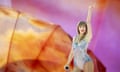 Taylor Swift, a tall white woman with blond hair and bangs, wears an iridescent body suit on a stage with an orange and purple background.