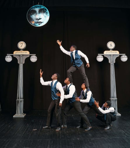 Five black acrobats in formal attire, one standing on the shoulders of two who are among four people making a sloping shape collectively