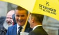 Angus MacNeil: he wears a blue suit and is standing laughing under a large yellow umbrella which bears the slogan Stronger for Scotland
