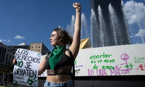 A woman with a green neckerchief stands with one arm raised and the other holding a poster with a Spanish slogan in support of abortion.