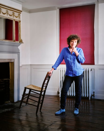 Jeanette Winterson wearing a blue shirt, standing in renovated 18-century living room with a fireplace, leaning on a chair