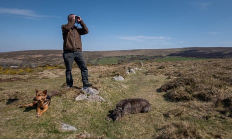 A man stands on the remains of a wall in a rugged green landscape with two dogs lying at his feet