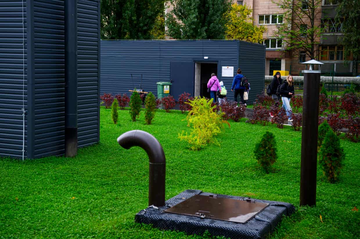 People leave an underground school’s entrance building, crossing a lawn with shrubs