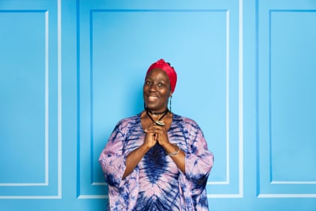 Pauline Tomlin is photographed smiling and wearing a red head scarf and tie-dyed top standing in front of a blue background