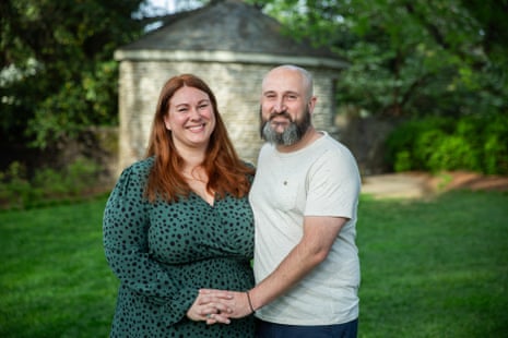 Lili and Luke standing close together smiling at the camera, Lili's right hand clasped in Luke's left hand, on a large lawn with a small building behind them