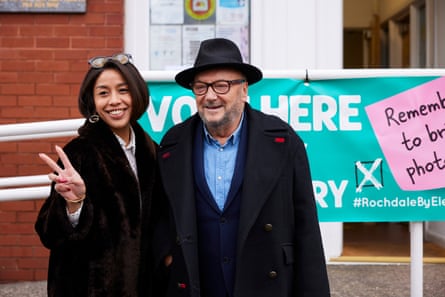 Galloway and Pertiwi stand in front of a polling station smiling. He wears a wide-brimmed black felt hat and black-rimmed glasses, a pale blue open-necked shirt and a heavy black overcoat with wide lapels and a red buttonhole detail. He is stocky with a trimmed grey beard. She appears to be significantly younger than him and has sleek black hair in a bob. She wears a brown fake-fur coat over a white blouse with black polka dots, plus sunglasses on the top of her head and a large earring in the shape of a G, possibly the Gucci symbol. She has long dark red fingernails and is making a v for victory sign.