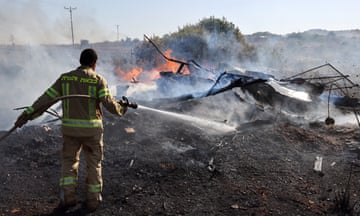 An Israeli firefighter puts out flames in a field: he wears overalls and is using a hose to douse flames among the wreckage of buildings; a hillside with small trees and power lines is seen in the background
