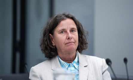 Woman in pale suit with light blue check shirt sitting beside microphones