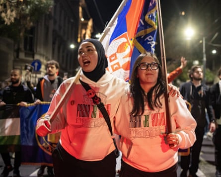 Two young women in matching white hooded sweatshirts sing and smile as they carry flags through a night-lit street