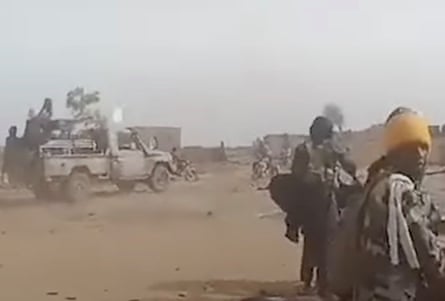 Blurred image of two men, one in military apparel and a yellow headband, in an arid landscape with a vehicle in the distance.