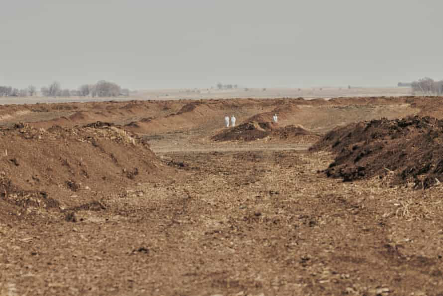 Two figures in white protective suits are seen in the distance amid mounds of bare soil 