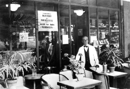 A man wearing a bow-tie and white overcoat stands outside his restaurant.