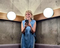A woman with a blue dress smiles while standing up holding her phone in a restaurant