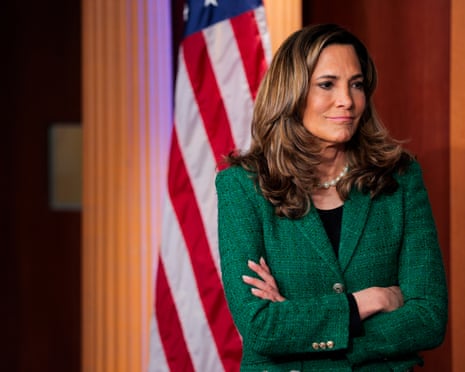 a woman in a green blazer stands with her arms crossed, an american flag behind her