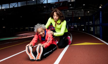 Ida Keeling sitting on a race track in kit, bending forward and holding the toes of her trainers, with her daughter behind her, kneeling and with her hands on her mum's shoulders