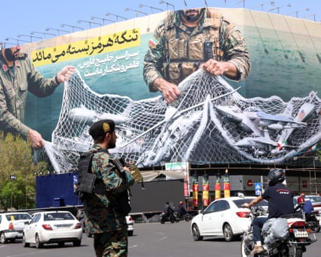 A man in military uniform stands as cars and motorcycles pass. The billboard in the background shows military personnel holding a net full of planes and ships