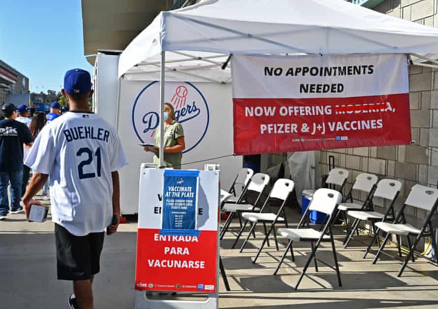 Un bărbat într-o pălărie și tricou din Los Angeles Dodgers trece pe lângă un cort cu semne care oferă bilete gratuite pentru fiecare vaccin luat înainte de joc.