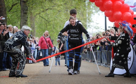 Claire Lomas crosses the finishing line held by a Cockney pearly king and queen. She is wearing a bionic suit and on crutches.