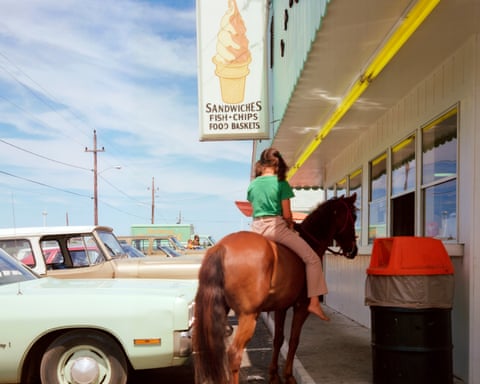 A girl on horseback approaches a drive-through window at a roadside ice cream stand with vintage cars parked outside