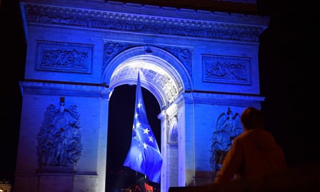 People gather at the Arc de Triomphe as it is lit up in blue alongside the EU flag