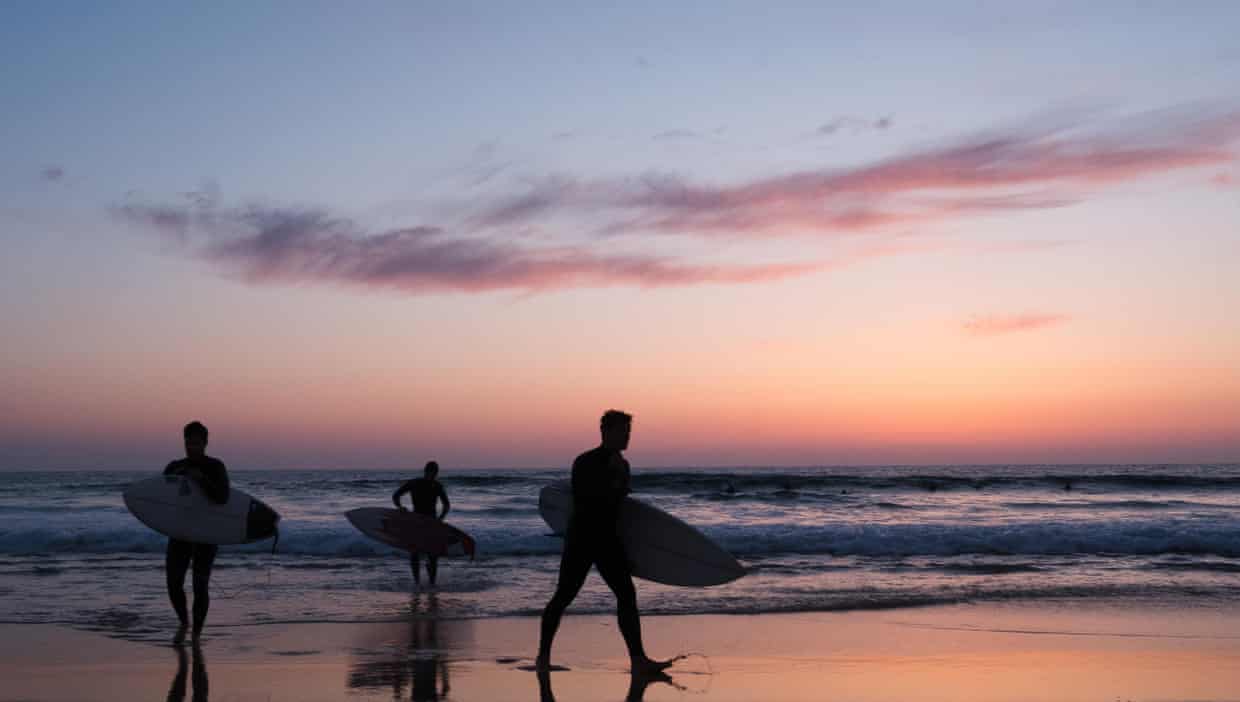 Three surfers carry their boards on the beach at sunset